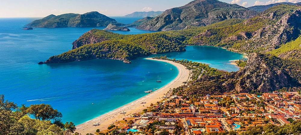 A crescent sandy beach curves along a turquoise bay, with green hills and a town of red-roofed buildings at the shore.