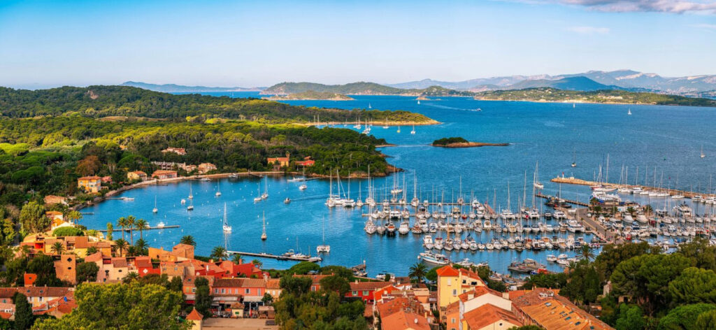 A coastal harbor with numerous sailboats and orange-roofed houses along the shore, backed by green hills and blue water.