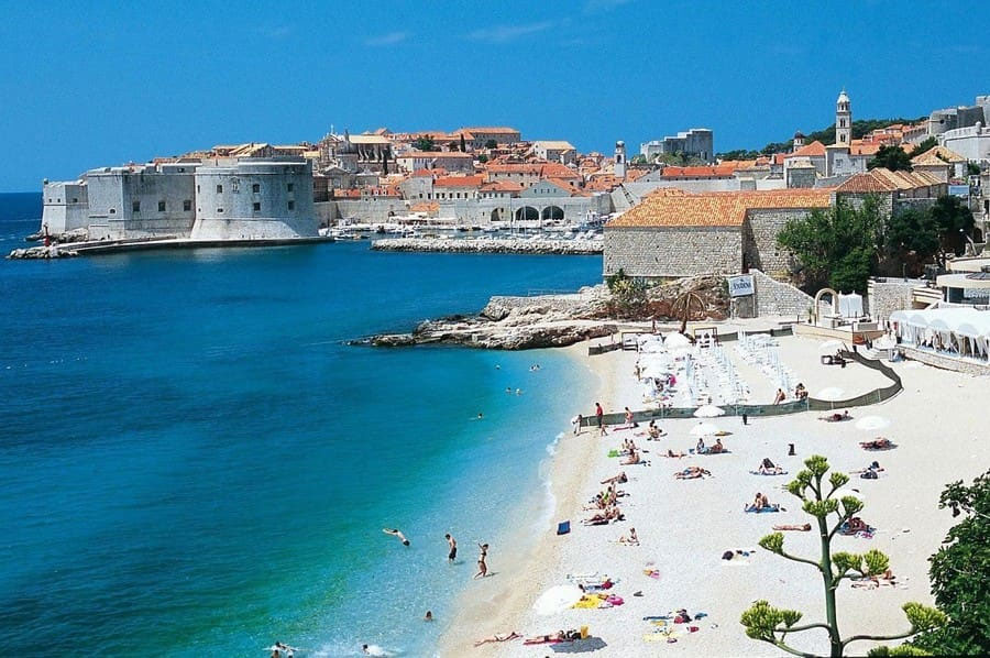 Sunny Adriatic beach with turquoise water and a historic stone city wall in the background; people sunbathing on the white sand.