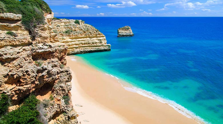 Sunlit beach with beige cliffs, golden sand, and turquoise water along a rocky coastline in clear weather