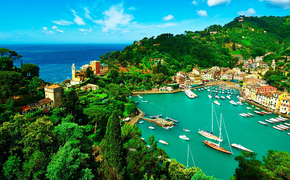 Coastal Italian harbor with colorful buildings, boats in a turquoise bay, and green hillside scenery under a bright blue sky.