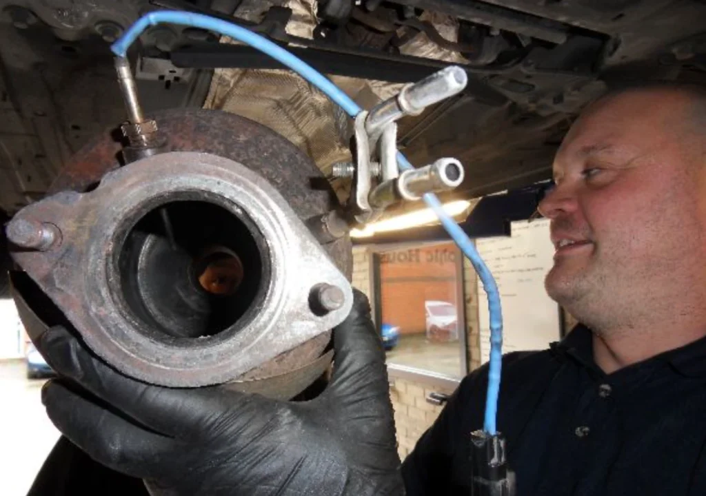Mechanic wearing gloves inspects a rusted brake rotor and hub with blue hydraulic line attached under a lifted vehicle.