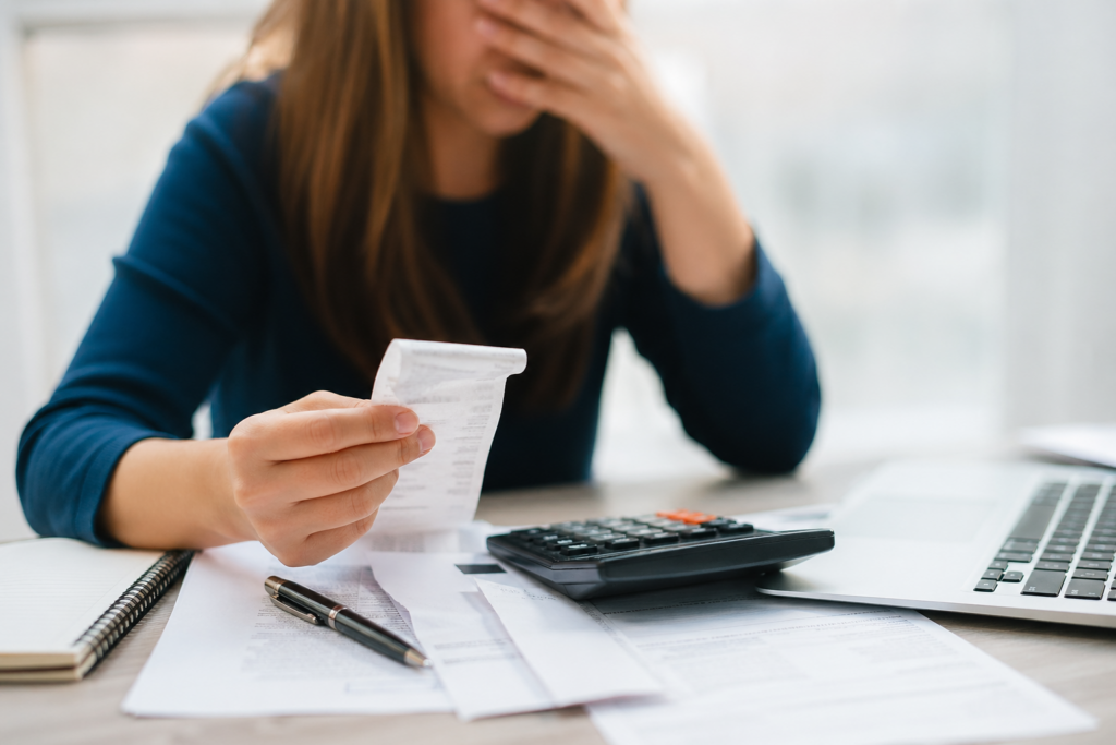 Woman sits at a desk, distressed, holding a receipt with a calculator and papers nearby with a laptop on the desk.