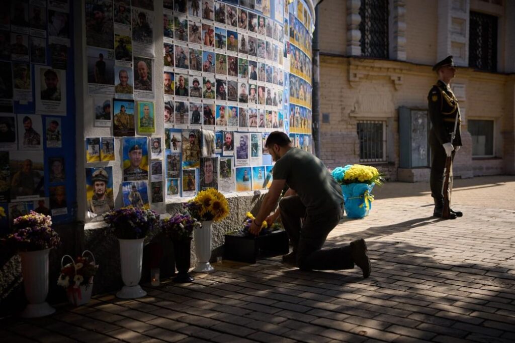 Man kneels to place flowers at a memorial wall covered with portrait posters, with a guard standing nearby.