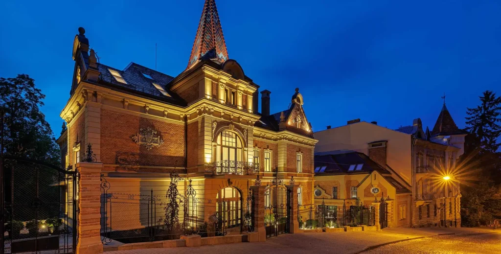 Night view of an ornate brick mansion illuminated by warm lights, with a tall conical tower and iron gates against a deep blue sky.