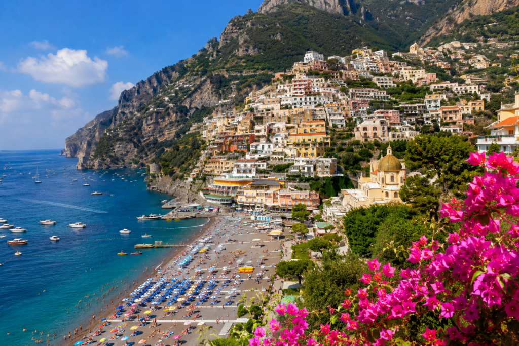 Colorful cliffside town along a blue sea with a crowded pebble beach and boats, pink flowers in the foreground.