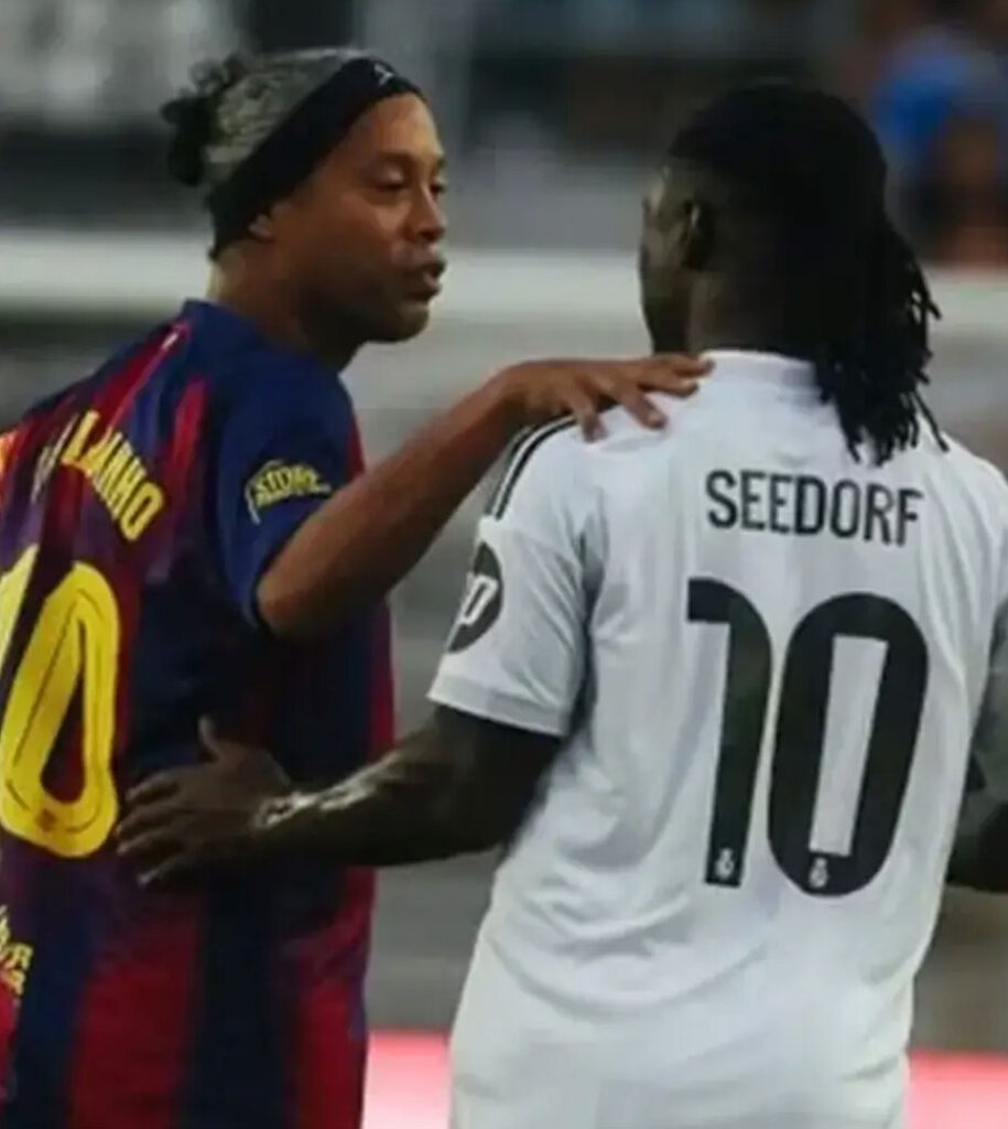 Two soccer players greet each other on the field, one in a white Seedorf jersey and the other in a Barca kit, touching shoulders in a friendly exchange. Легенди Барселони і Реала зіграли в Майамі