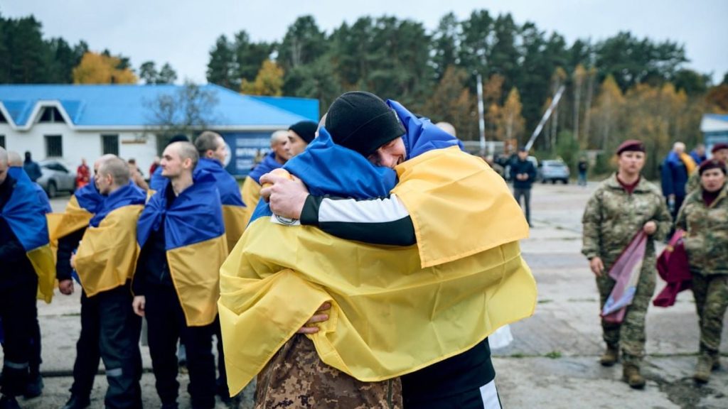 символічна сценична композиція: складений камуфляж, маленький прапор України й черевики на деревʼяному фоні