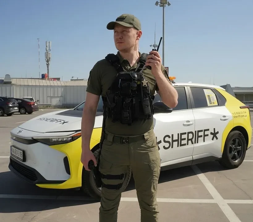 Охоронець із рацією біля службового автомобіля на відкритій парковці Фізична охорона об'єктів у Києві: пости охорони під ключ