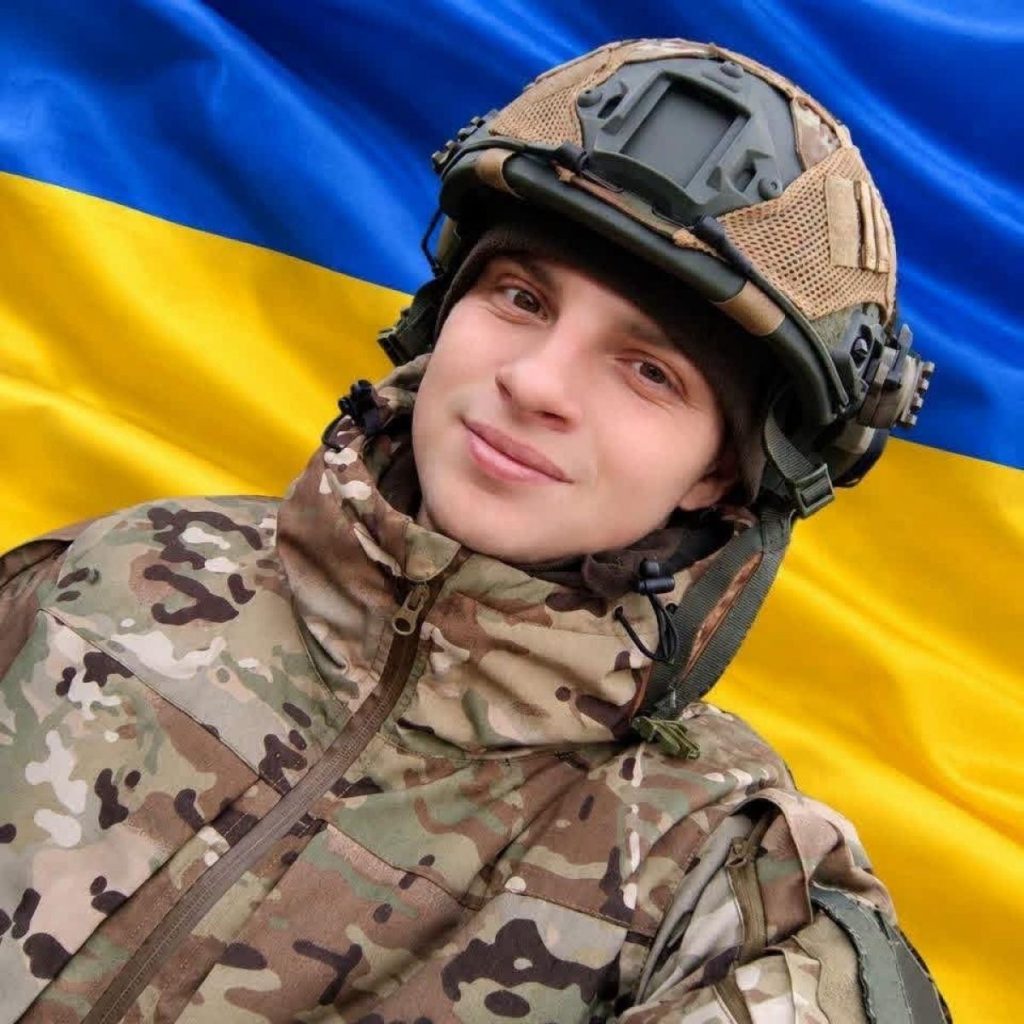 Складана військова каска на деревʼяному столі поруч зі жетонами та маленьким прапорцем України