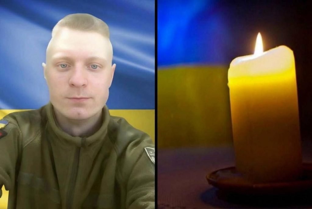 згорблений військовий шолом, свічка та маленький прапорець України на дерев'яному столі, помірне тепле освітлення
