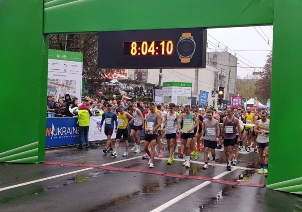 Бігові кросівки, стартовий номер і прапор України на столі, на фоні розмитий тепловізор та тактичні рукавички